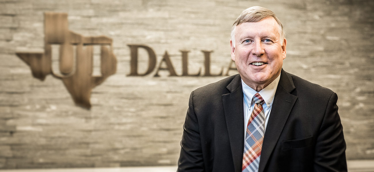 Benson smiles as he sits in front of a UT Dallas logo